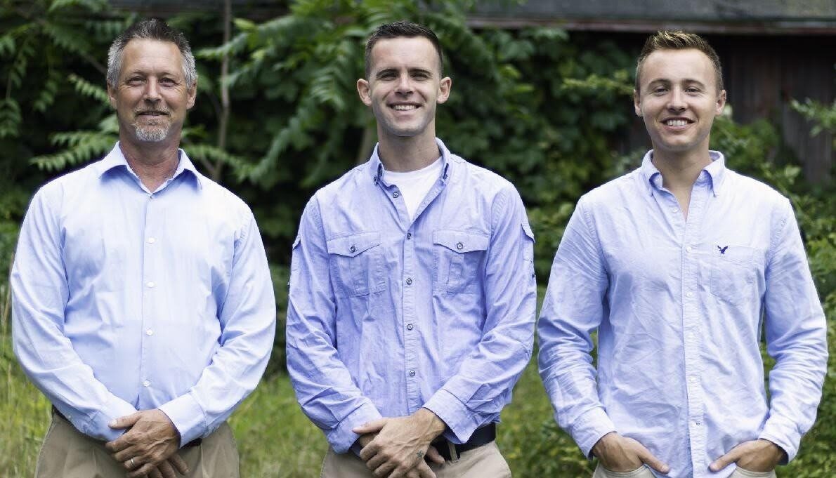 Three men smiling outdoors, dressed in light blue shirts.