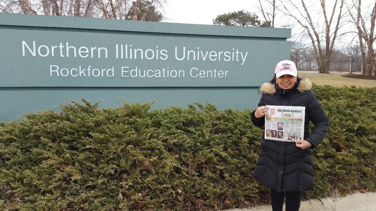 Person holding a newspaper in front of Northern Illinois University sign.