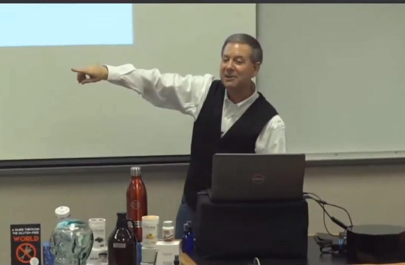 A man in a vest pointing while speaking in front of a laptop and bottles.