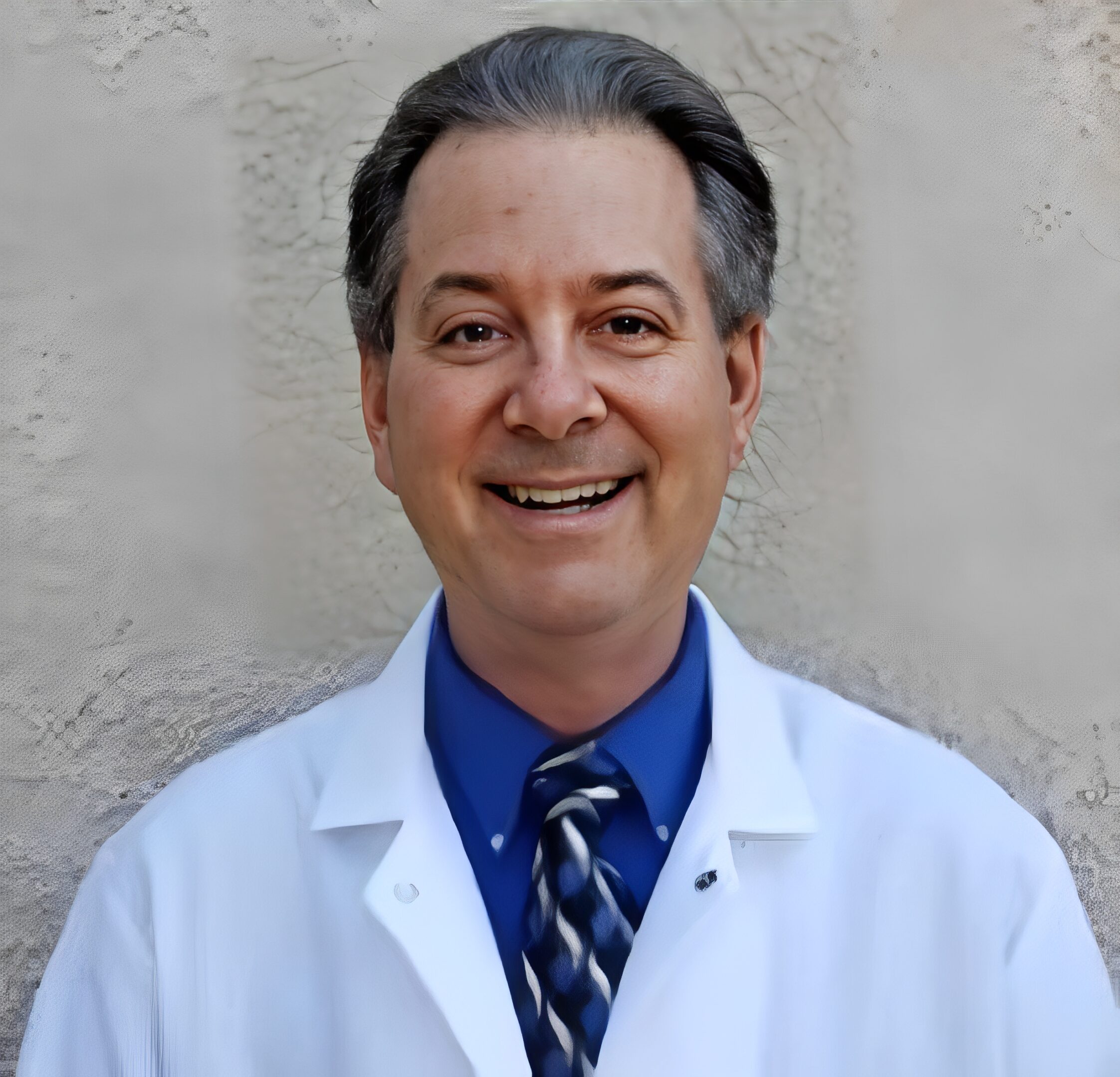 Smiling man in a white lab coat and tie against a plain background.