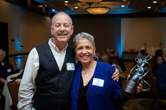 Two smiling people posing at an indoor event with name tags.
