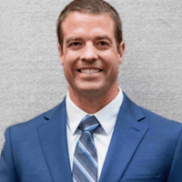 Smiling man in a blue suit and striped tie against a gray background.