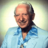 Smiling elderly man in a light blue jacket with a patterned shirt.
